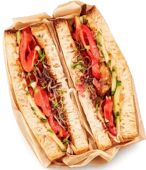Sandwich with toasted bread, tomato slices, leafy greens, cucumber, and sauce on parchment paper.