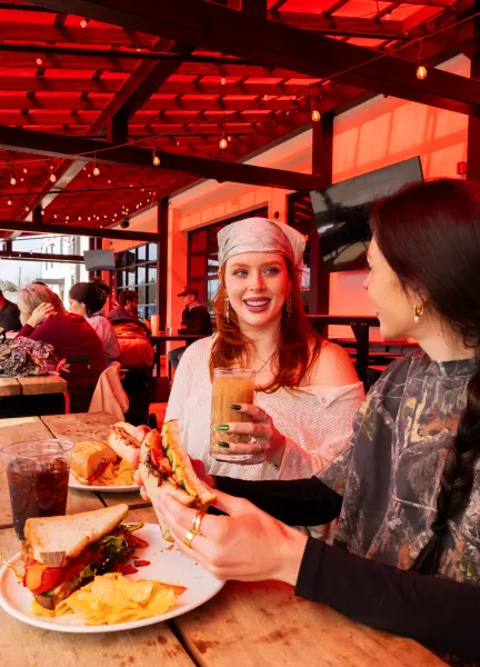 Two women smiling and enjoying sandwiches and coffee at a wooden table in a cozy restaurant.