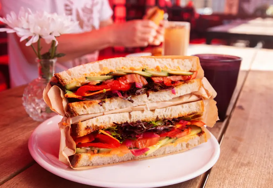 Fresh sandwich with turkey, lettuce, tomato, and cucumber on white plate on wooden table.