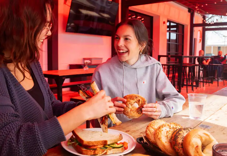Two women sitting on the sunbeam patio eating bagels and sandwiches