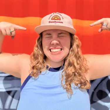 Woman smiling with eyes closed pointing at her beige Sunbeam brand hat standing against a red wall.