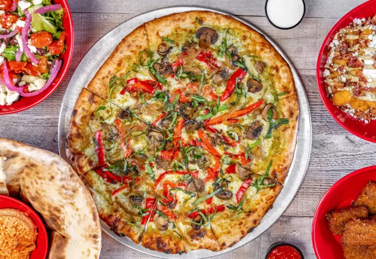 Vegetable pizza with mushrooms, red pepper, and greens on a metal tray among side dishes.