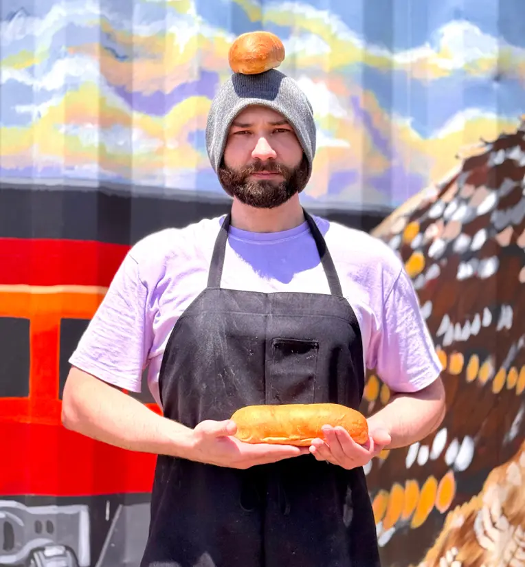 Man wearing gray beanie and black apron holding bread loaf and balancing a bun on his head.