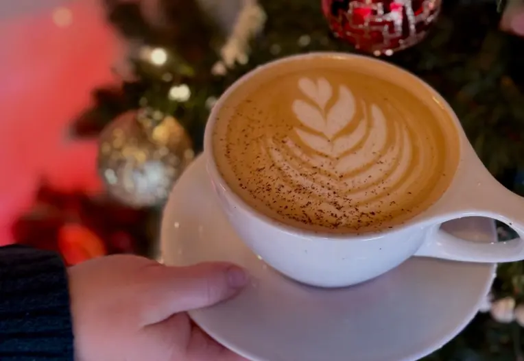Hand holding a white cup of latte with leaf latte art and spice on top near a decorated Christmas tree.