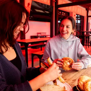 Two women sitting on the patio of Sunbeam holding sandwiches and bagels