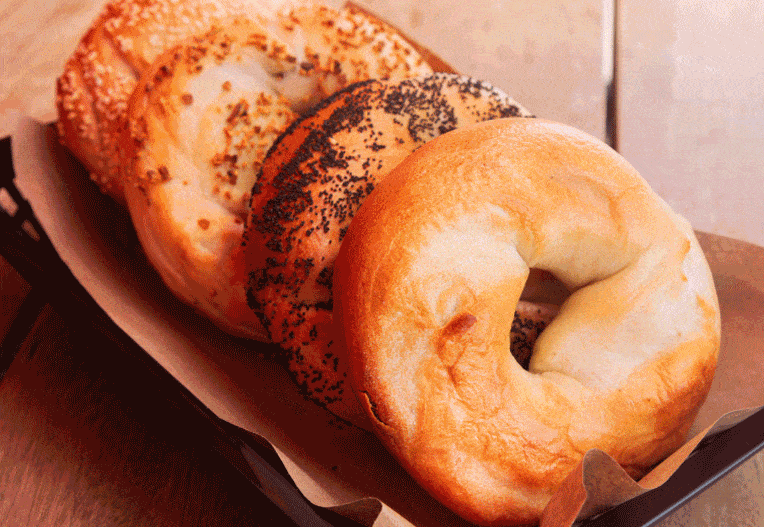 Gif including images of a tray of assorted bagels, a bagel sandwich wrapped in paper, chocolate chip cookies, someone holding a cheesy bagel sandwich, and baskets of bagels at the check out counter