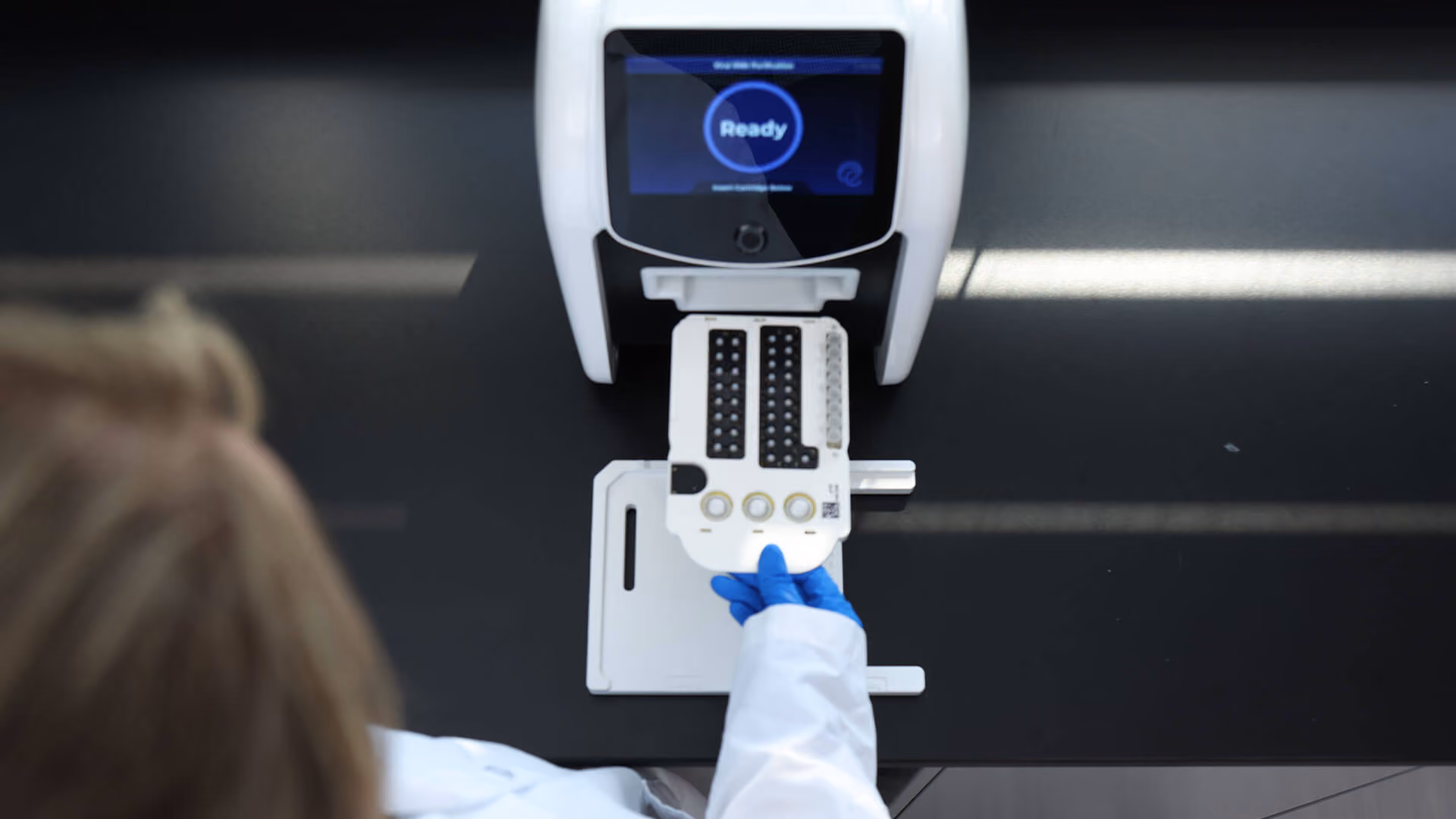 Person in lab coat and blue gloves inserting a white cartridge into a machine showing a ready screen.