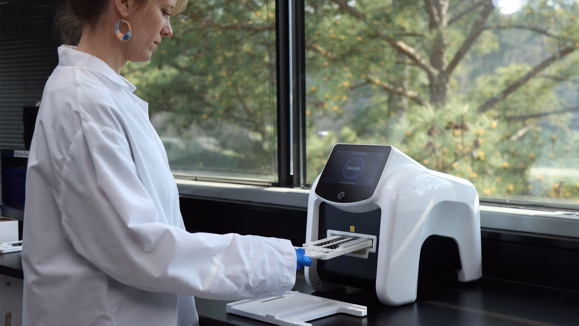 Lab technician in white coat loading a cartridge into a white diagnostic machine by a window.