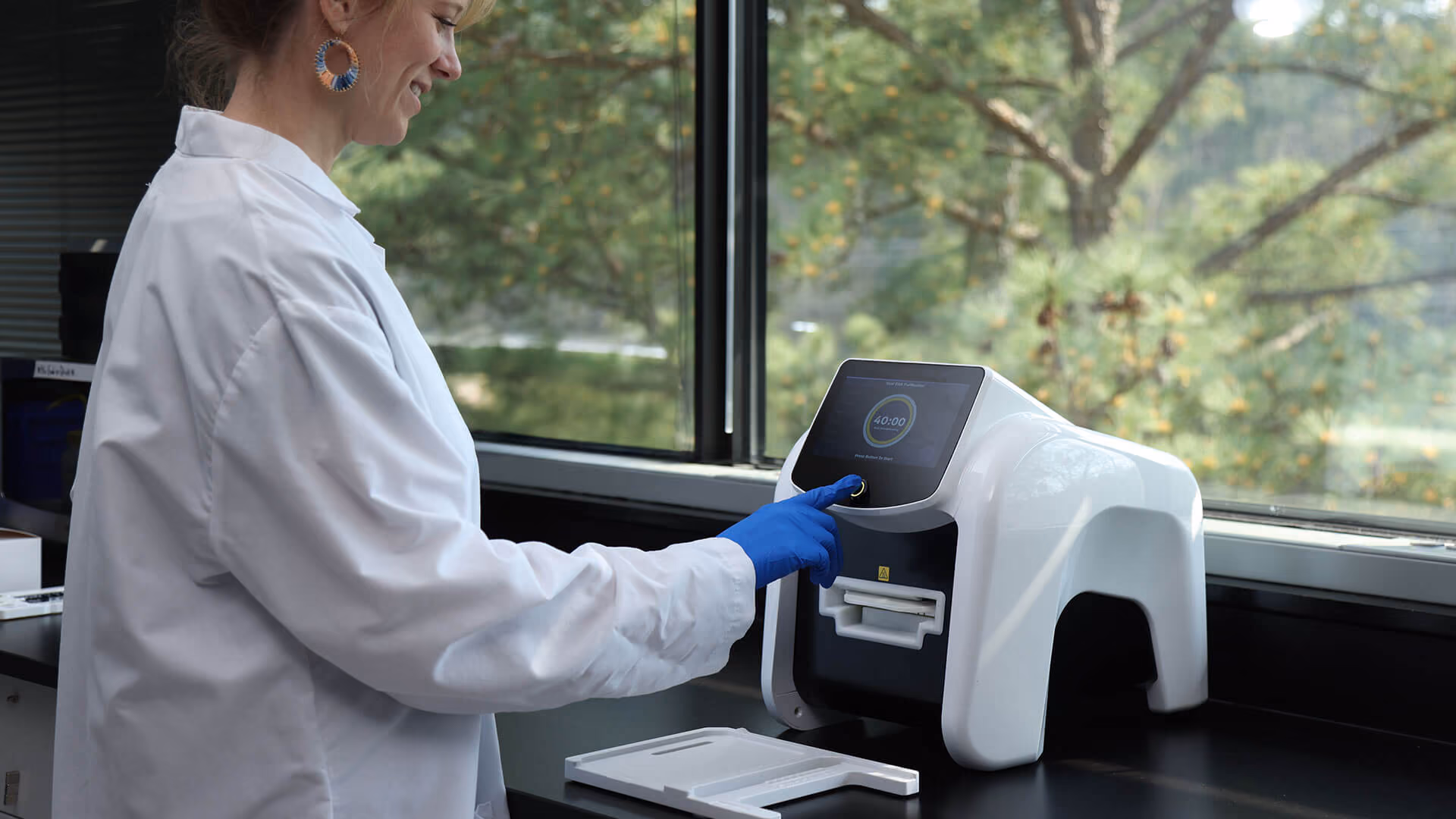 Person in lab coat and gloves pressing a button on a white scientific machine by a window.