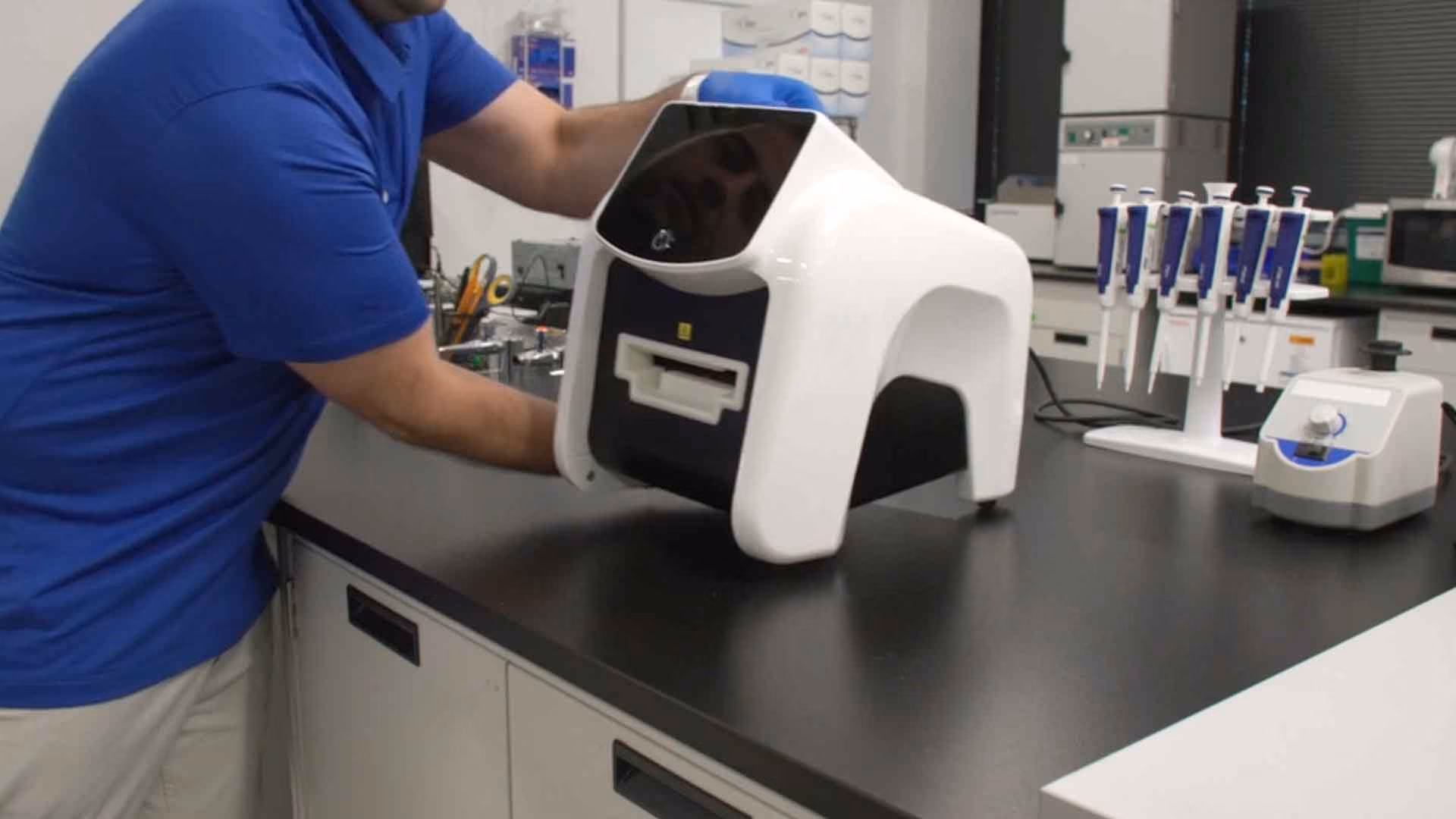 Person in blue shirt handling a white and black lab device on a bench with pipettes nearby.