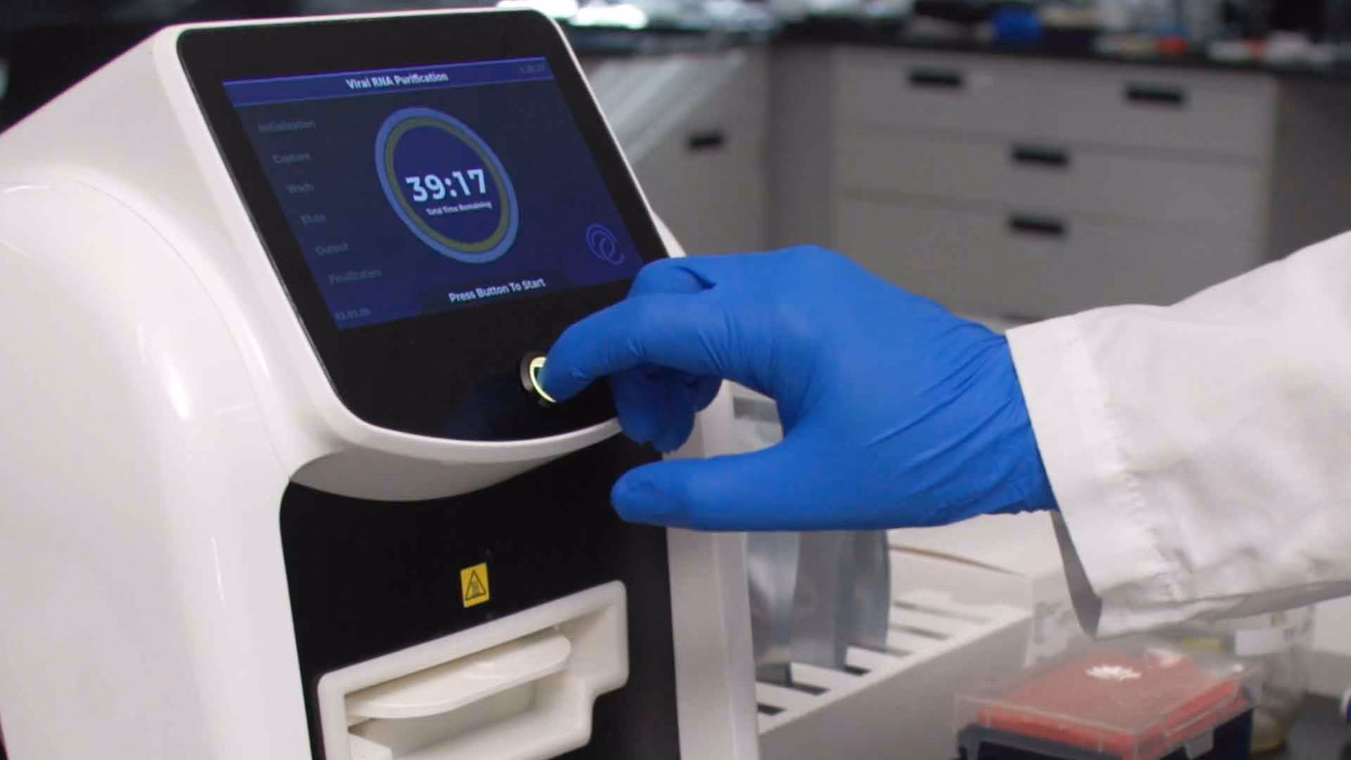 Hand in blue glove pressing a button on a white laboratory machine with screen showing viral RNA purification.