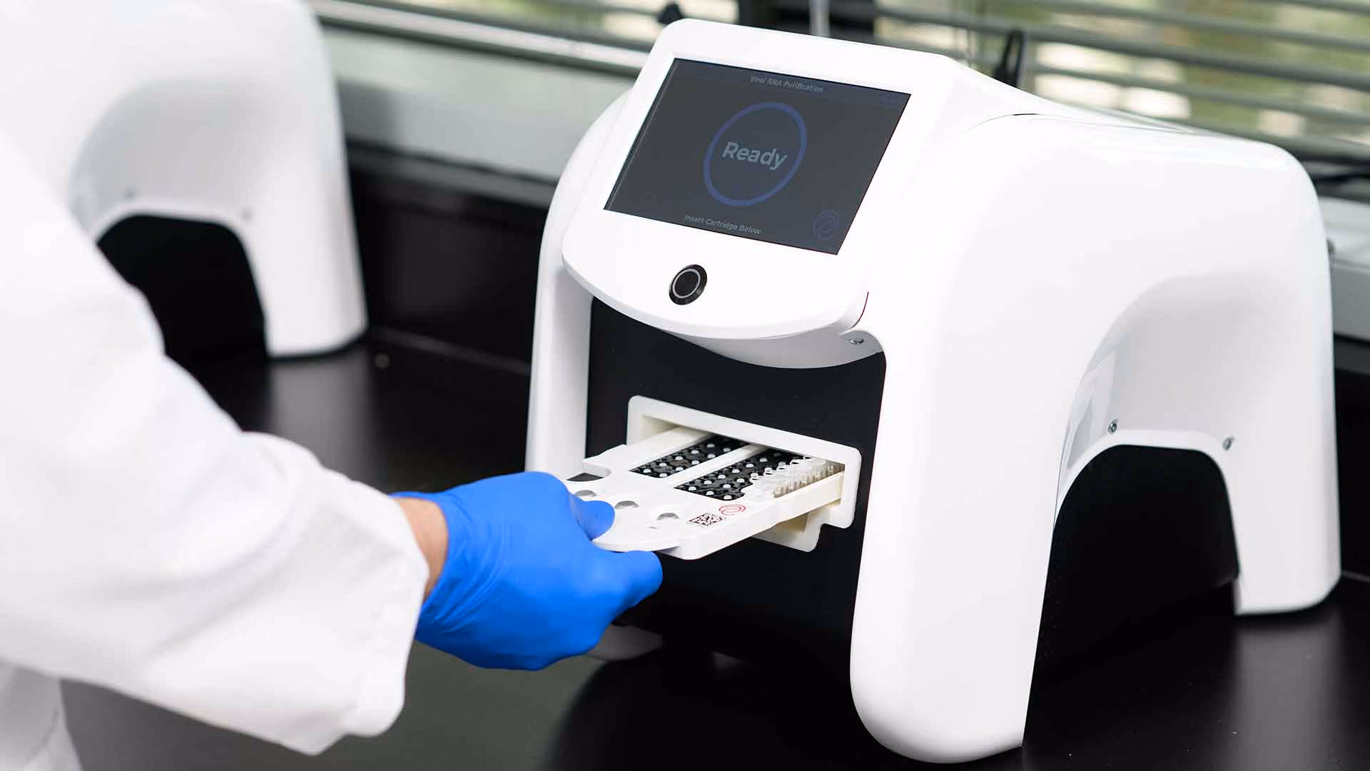 Hand in blue glove loading a cartridge tray into a white lab machine with a 'Ready' screen.