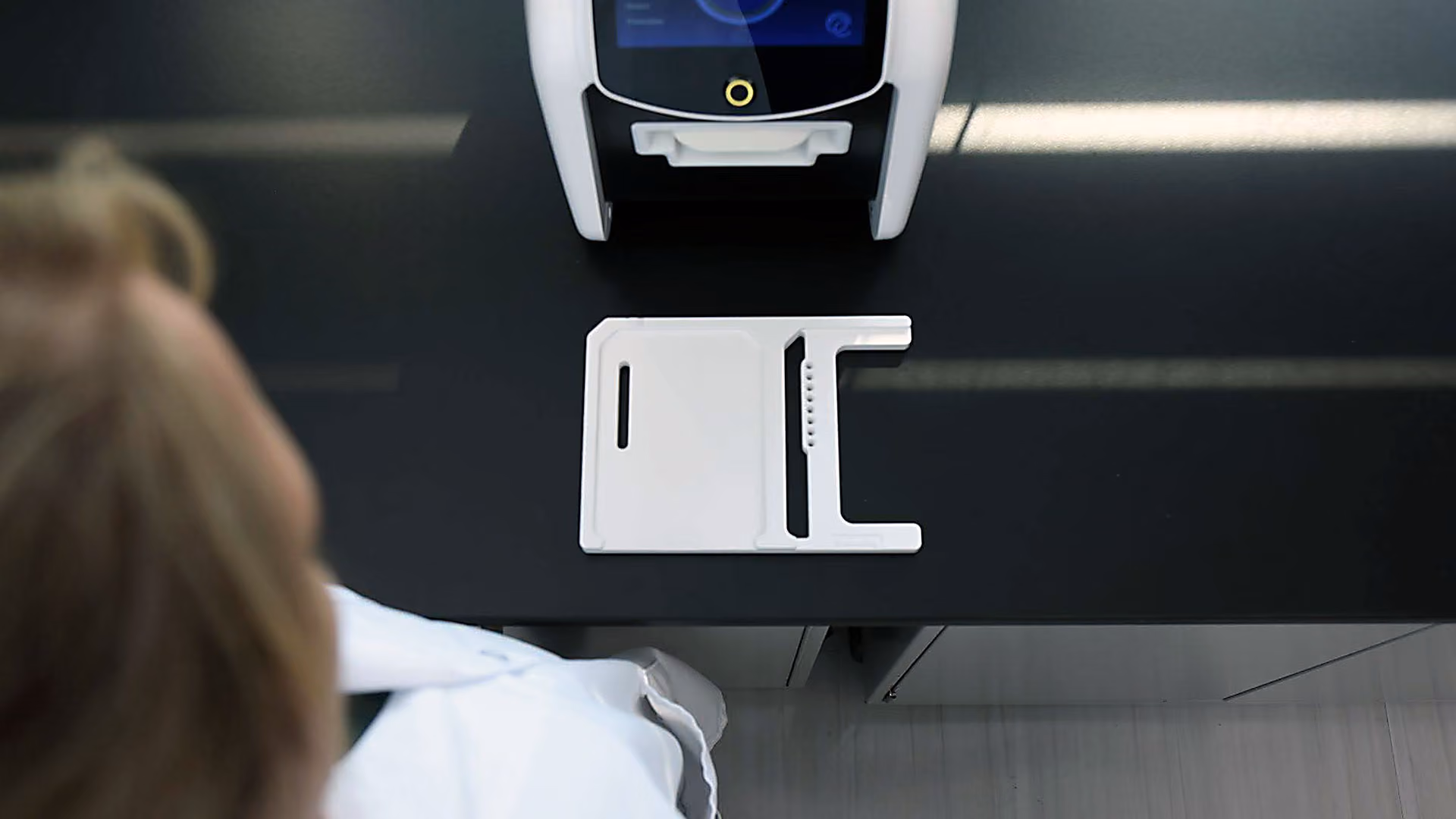 Top view of a white medical device on a black countertop with a blurred person in a white coat nearby.