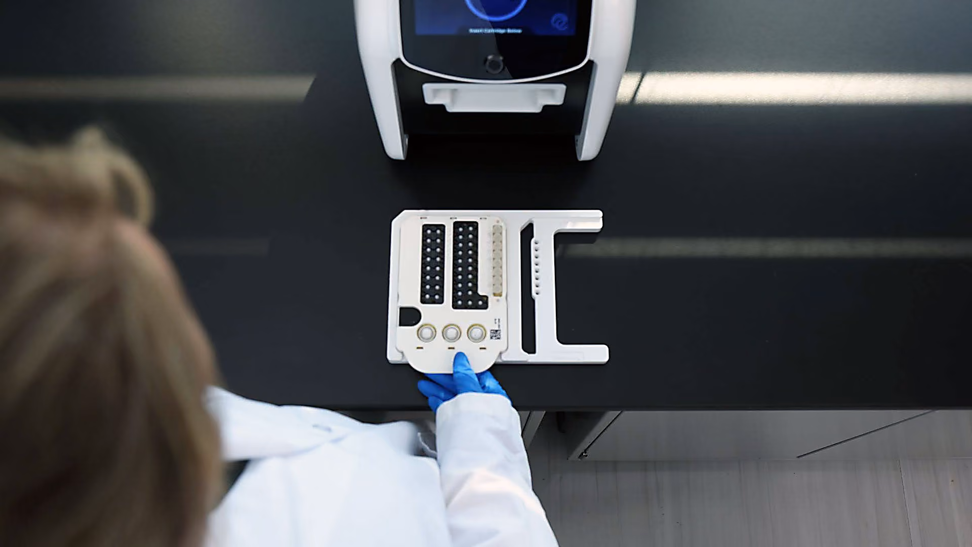 Overhead view of a lab technician wearing gloves placing a testing card on a black counter under a machine.