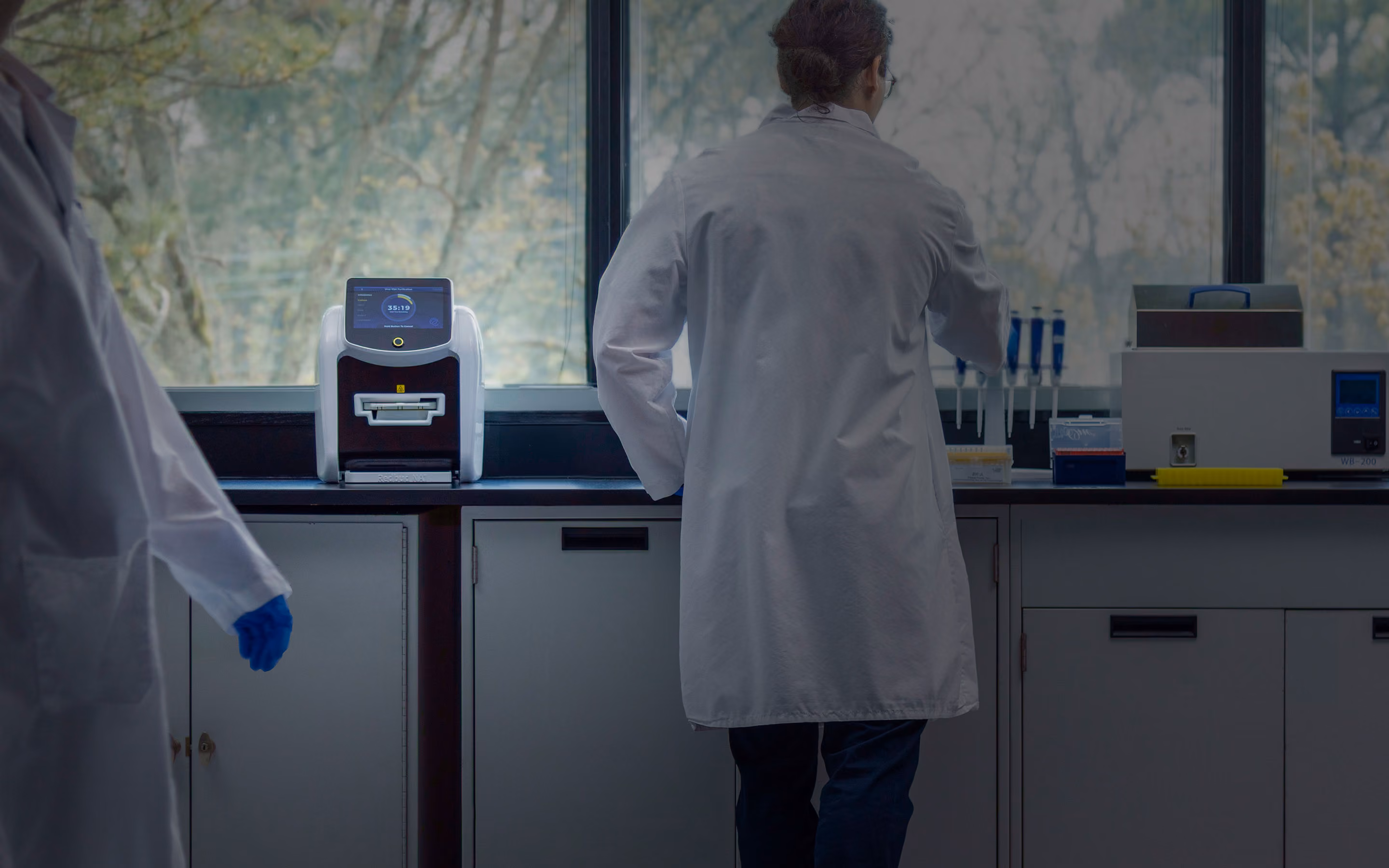 Scientist in a white lab coat working near lab instruments by a window with trees visible outside.