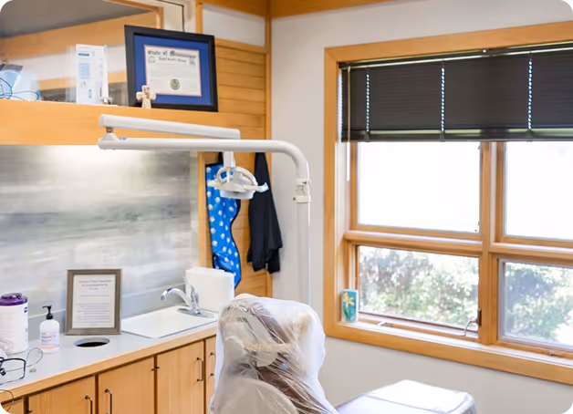 Dental exam chair covered in plastic beside a sink, cabinetry, and window with blinds in a clinic room.