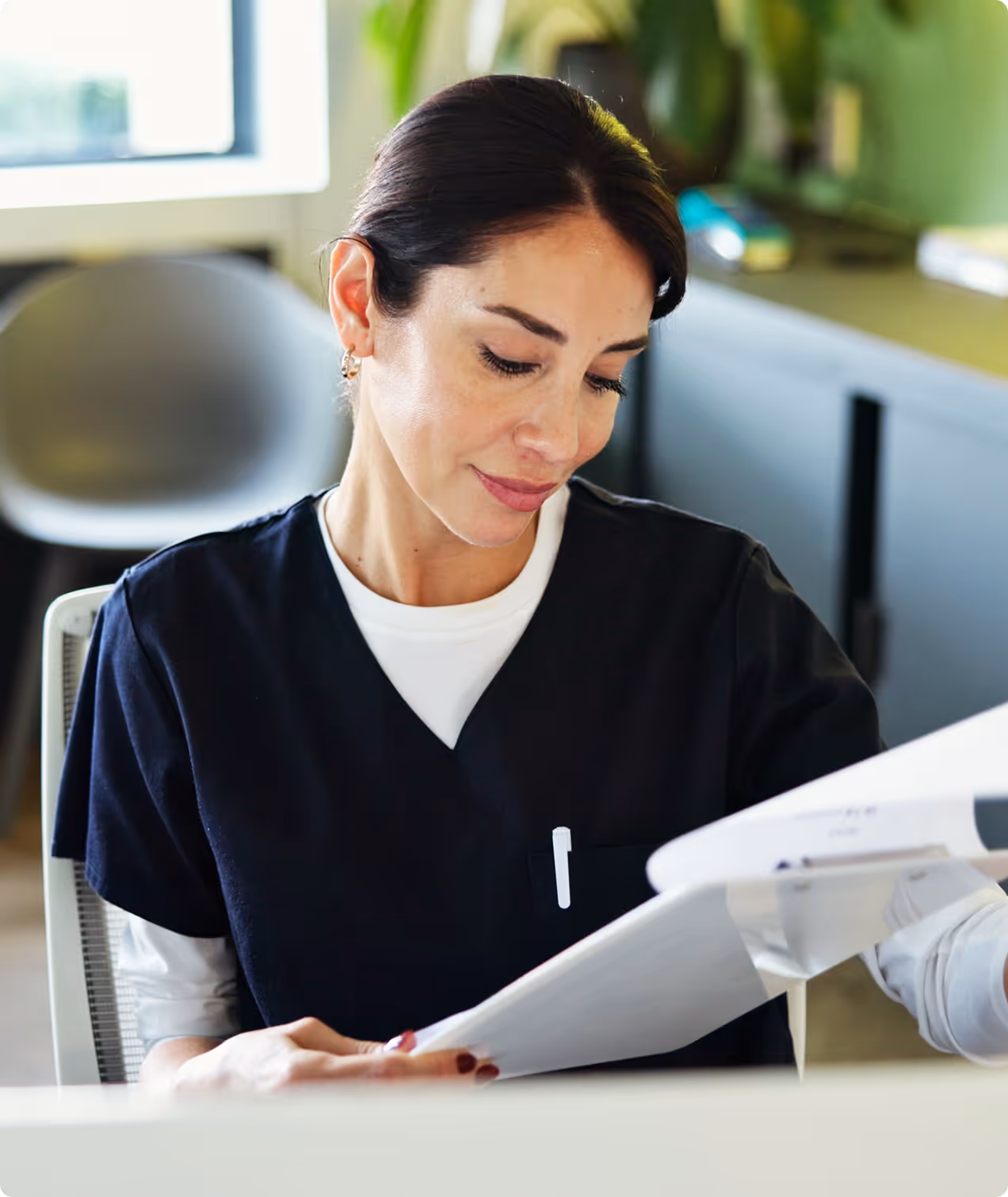 Woman in medical scrubs reviewing documents at an office desk with a chair and plants in background.