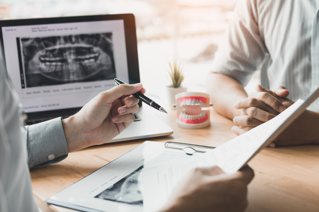 Person pointing to dental X-rays on laptop while discussing with patient at a desk with teeth model.