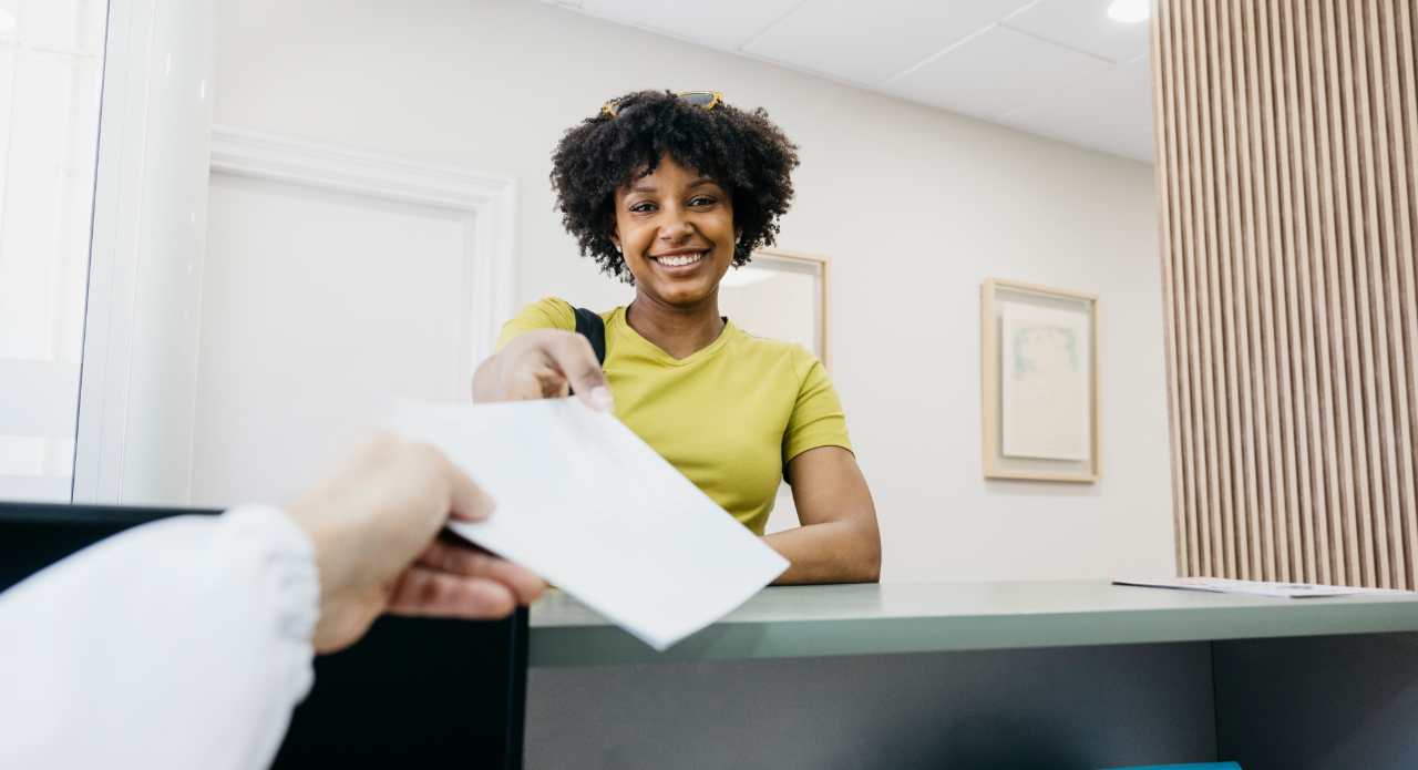 Smiling woman in green shirt handing a document to a person across a reception desk.