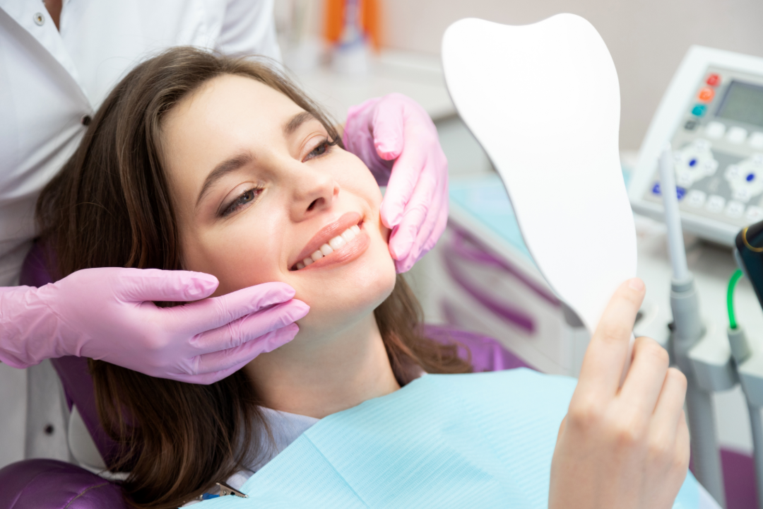 Woman smiles in dental chair as dentist wearing pink gloves examines her face with a tooth-shaped mirror nearby.