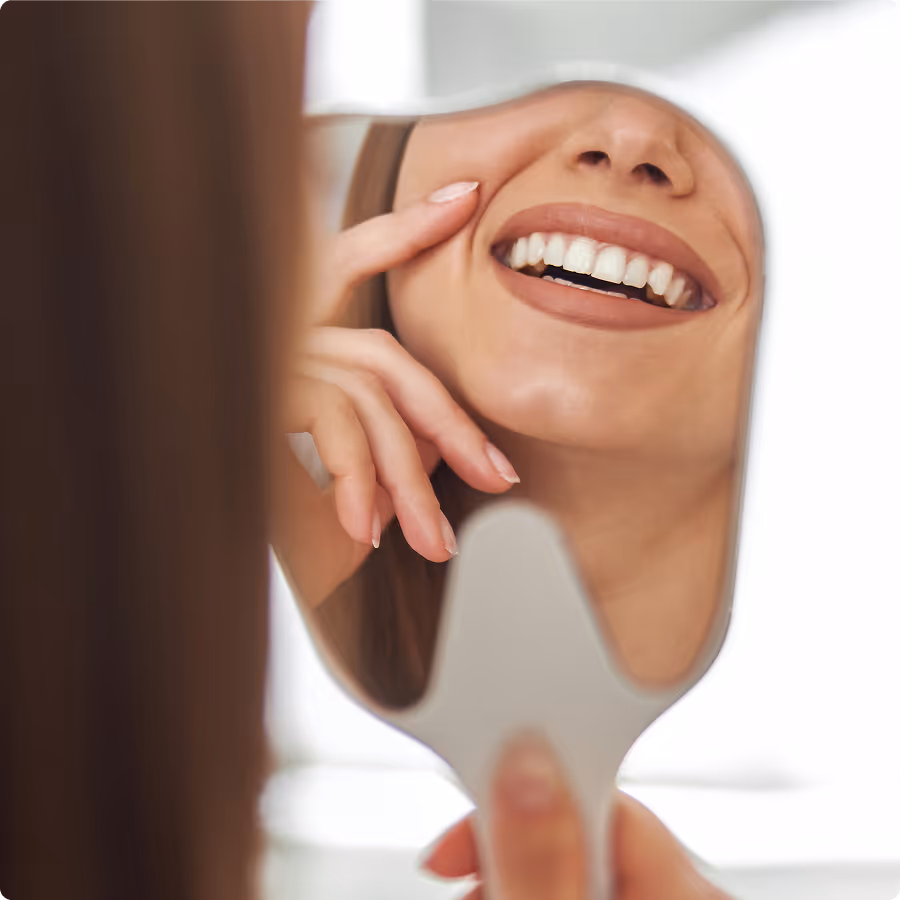 Close-up of woman smiling and touching cheek while looking at her reflection in a small handheld mirror.