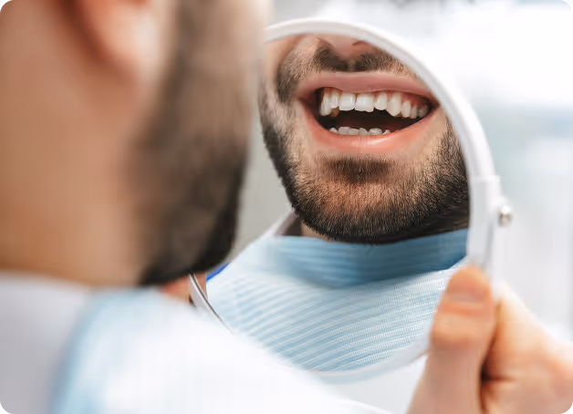 Bearded man holding a small mirror smiling, showing his teeth and dental bib.