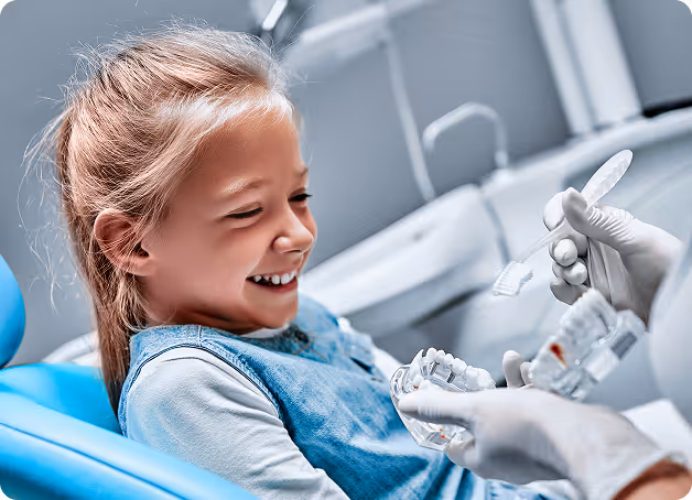 Smiling young girl sitting in dental chair while dentist shows dental model and toothbrush.