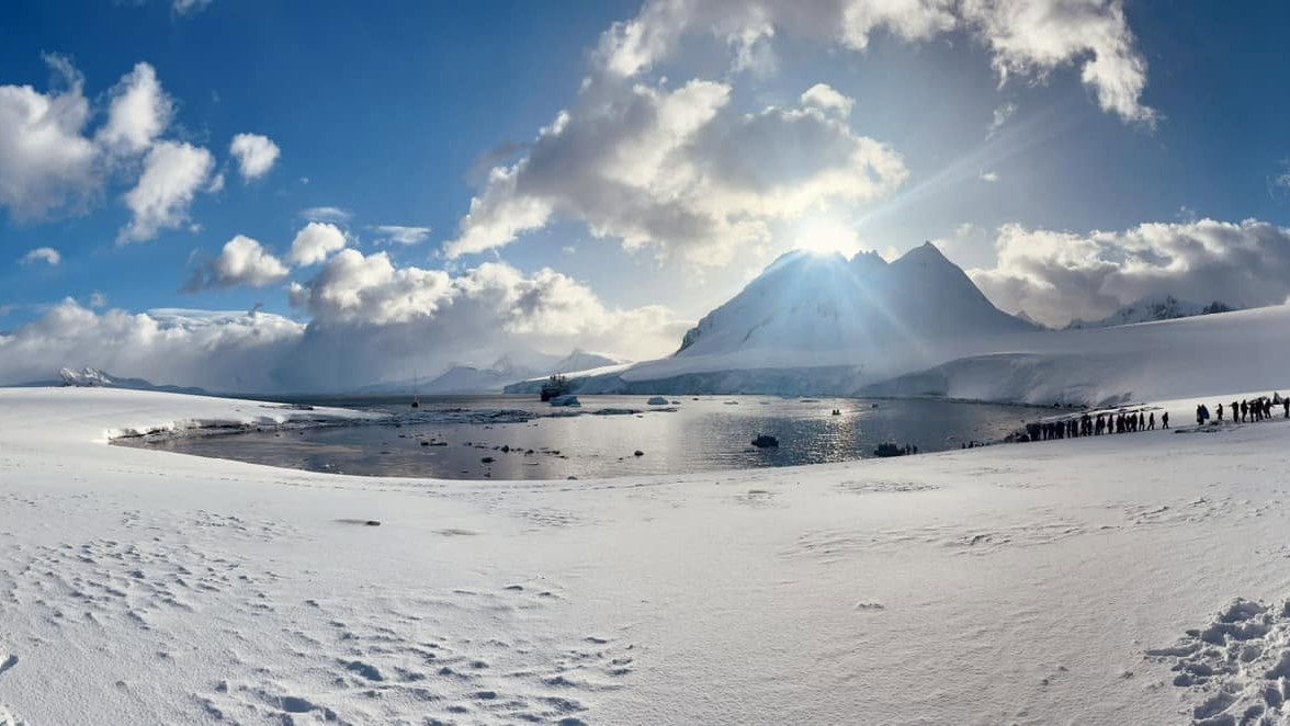 Sun bursting over a snow peak on King George Island, the ship at anchor in the bay and a line of people walking ashore across the snow