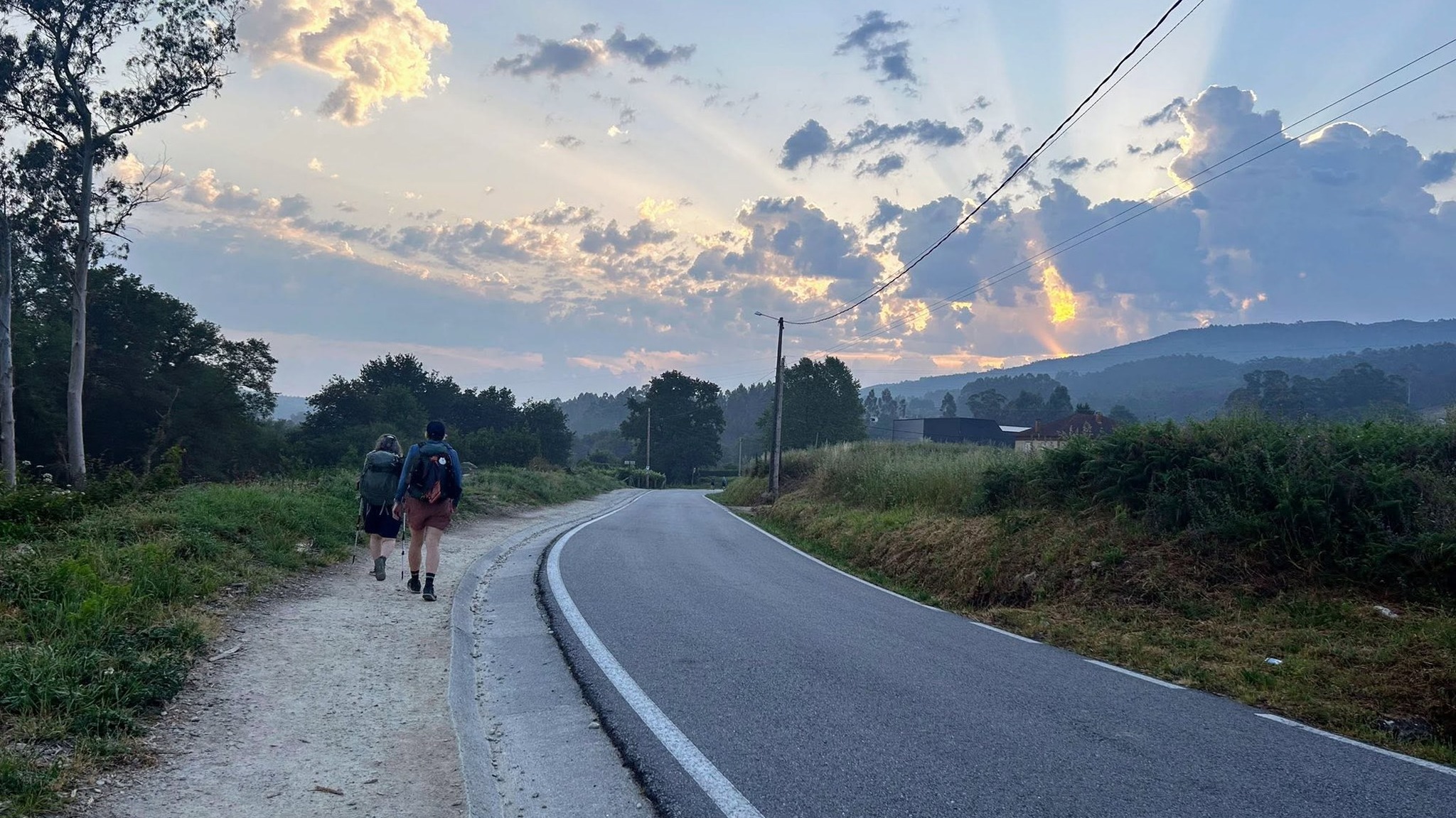 Two pilgrims with backpacks walking the gravel verge beside a curving country road at sunrise, shafts of light breaking through cloud over wooded hills ahead.