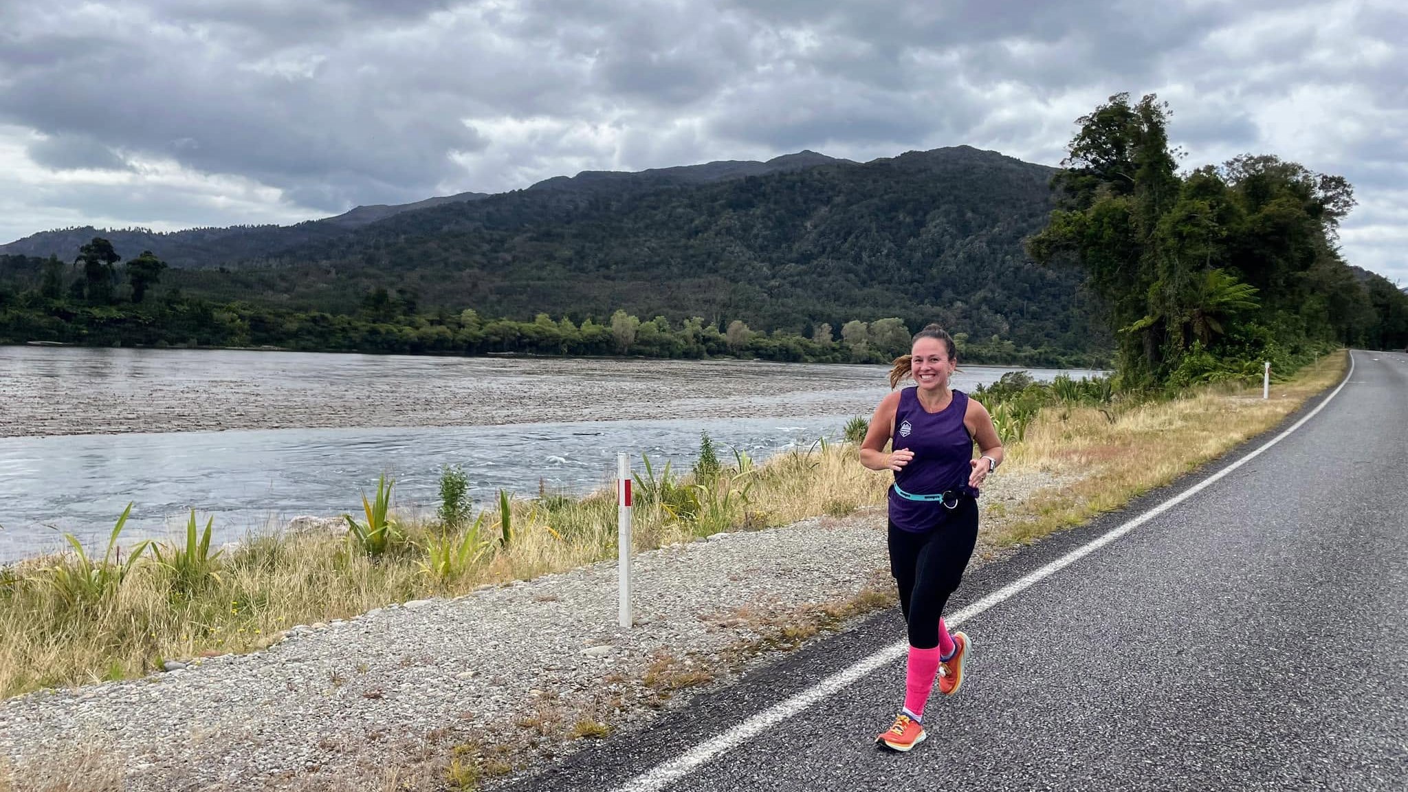 Nancy grinning mid-stride in a purple tank and pink compression socks on a road beside a wide braided river and forested hills