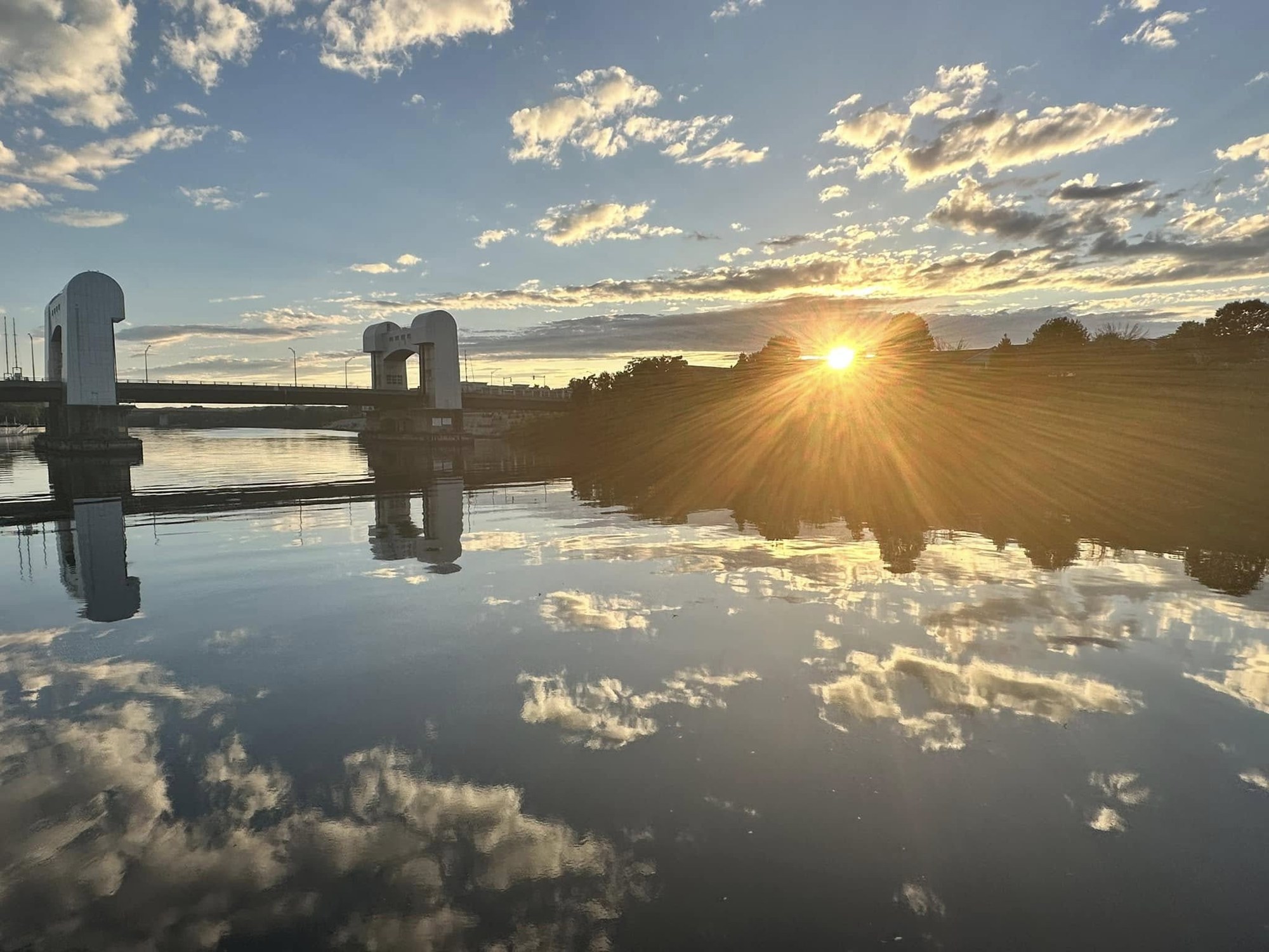 The sun setting behind trees beside a lift bridge, clouds mirrored in perfectly still water