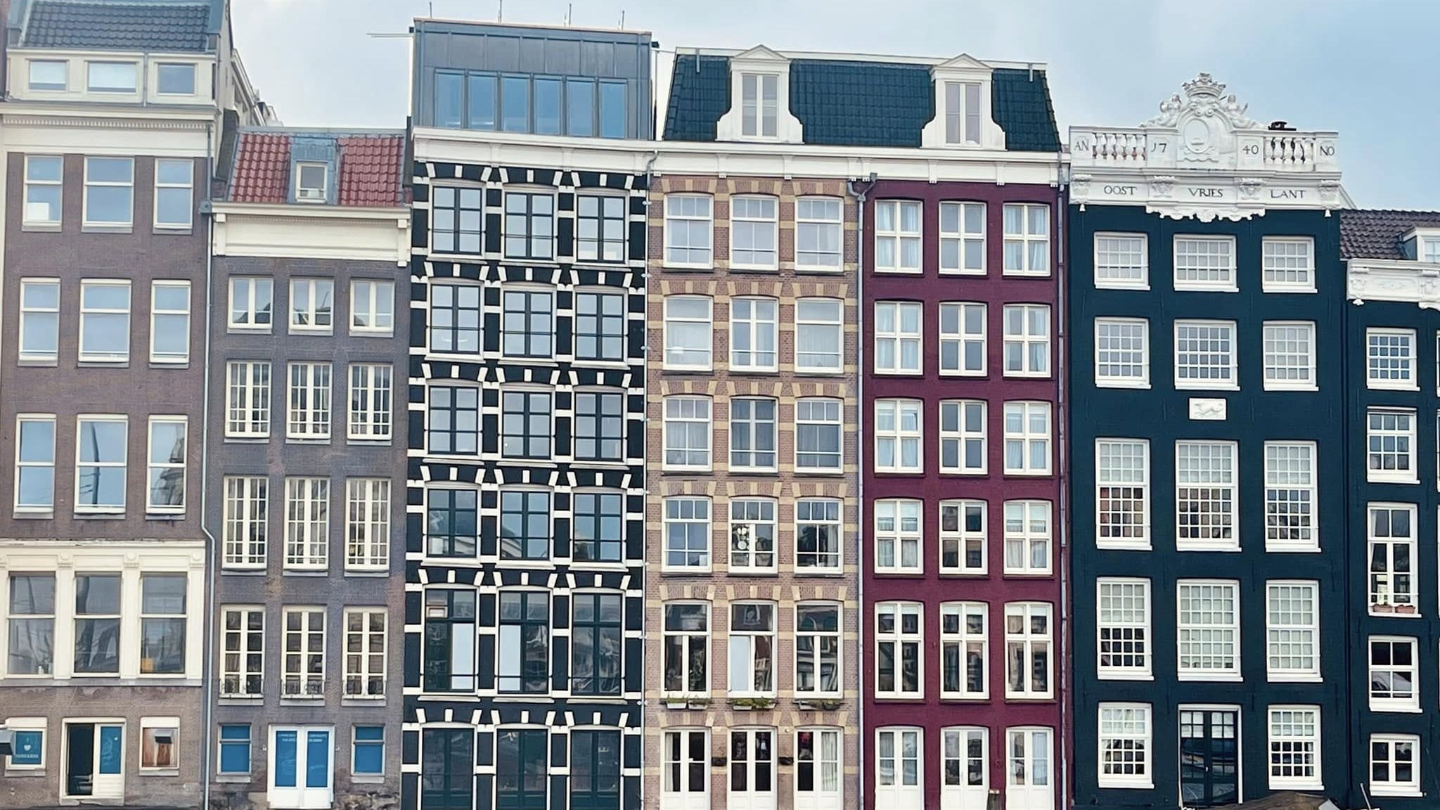 A row of tall, narrow canal houses on the Damrak in black, brick, maroon and dark green, leaning slightly against each other above the water