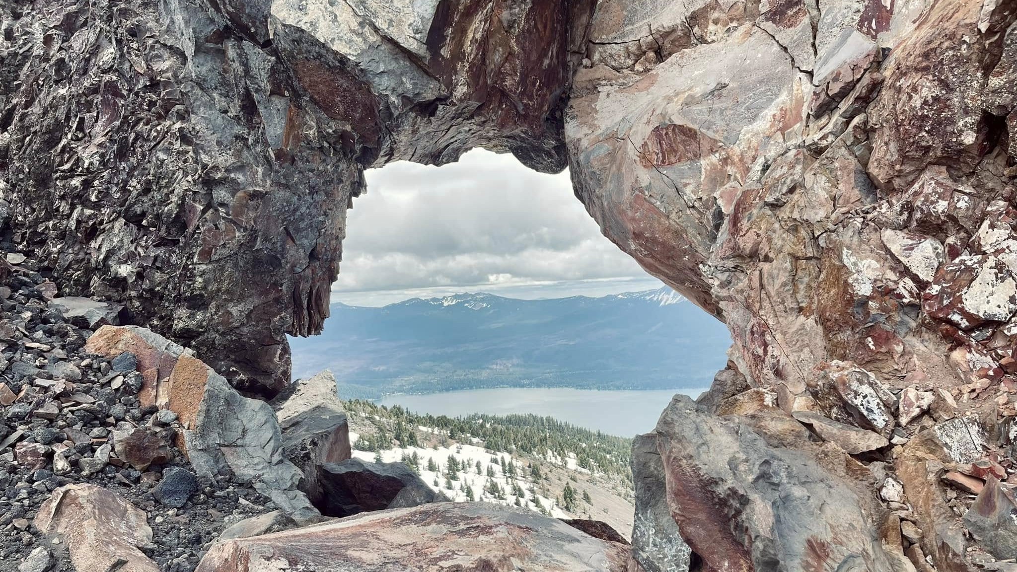 Through a natural rock window near the top of Mt. Bailey, a lake far below and snow-dusted ridges beyond