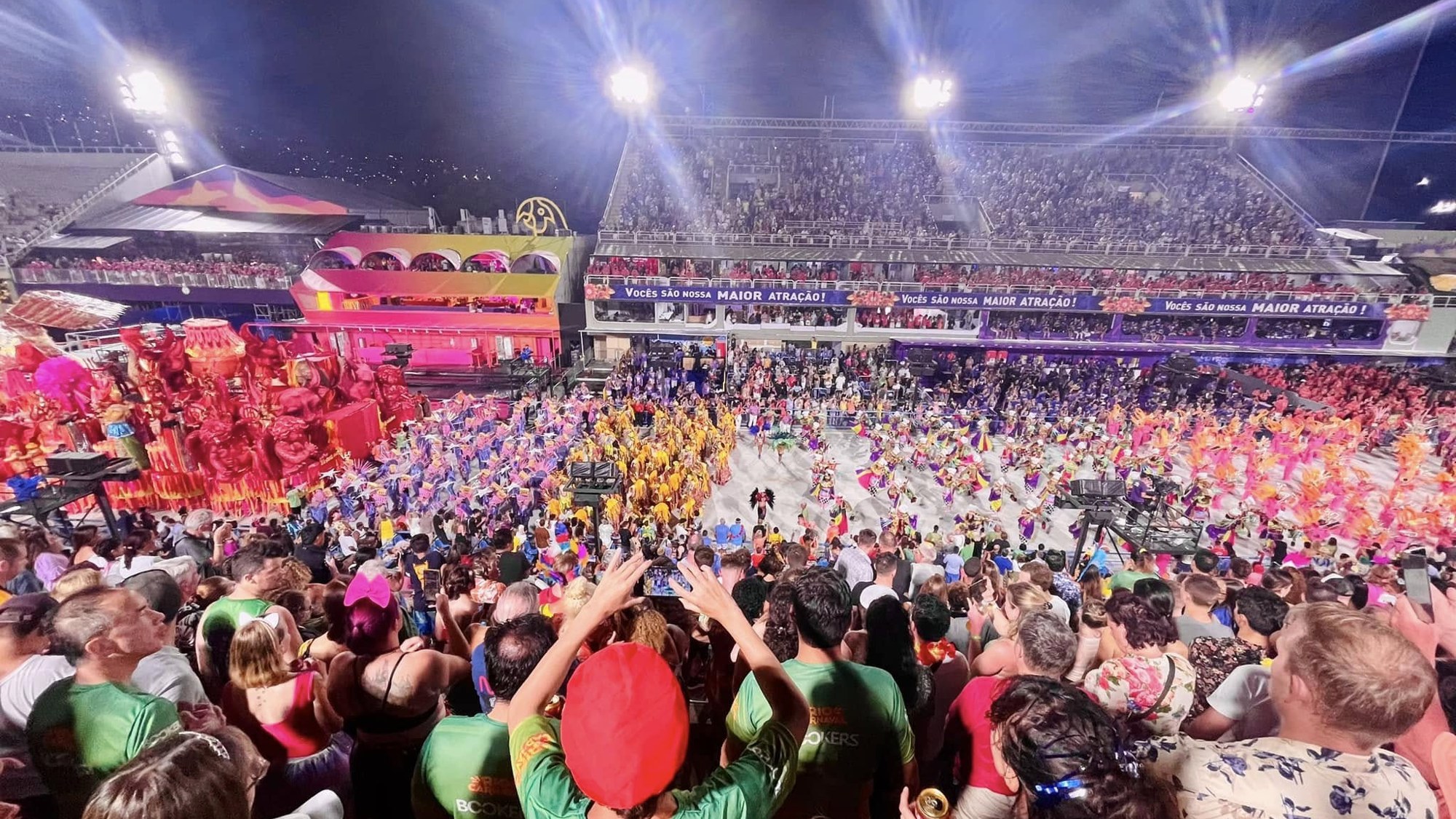 The Sambódromo at night from high in the stands, floodlights blazing over thousands of costumed dancers and a towering red float