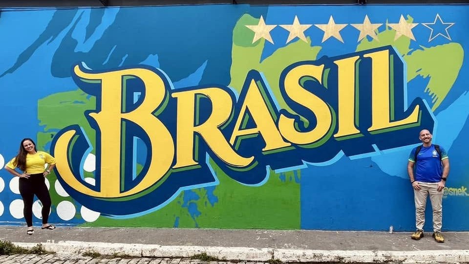 Nancy in a yellow Brazil shirt and Jay in the blue away shirt at either end of a giant BRASIL mural with five gold stars and an empty sixth