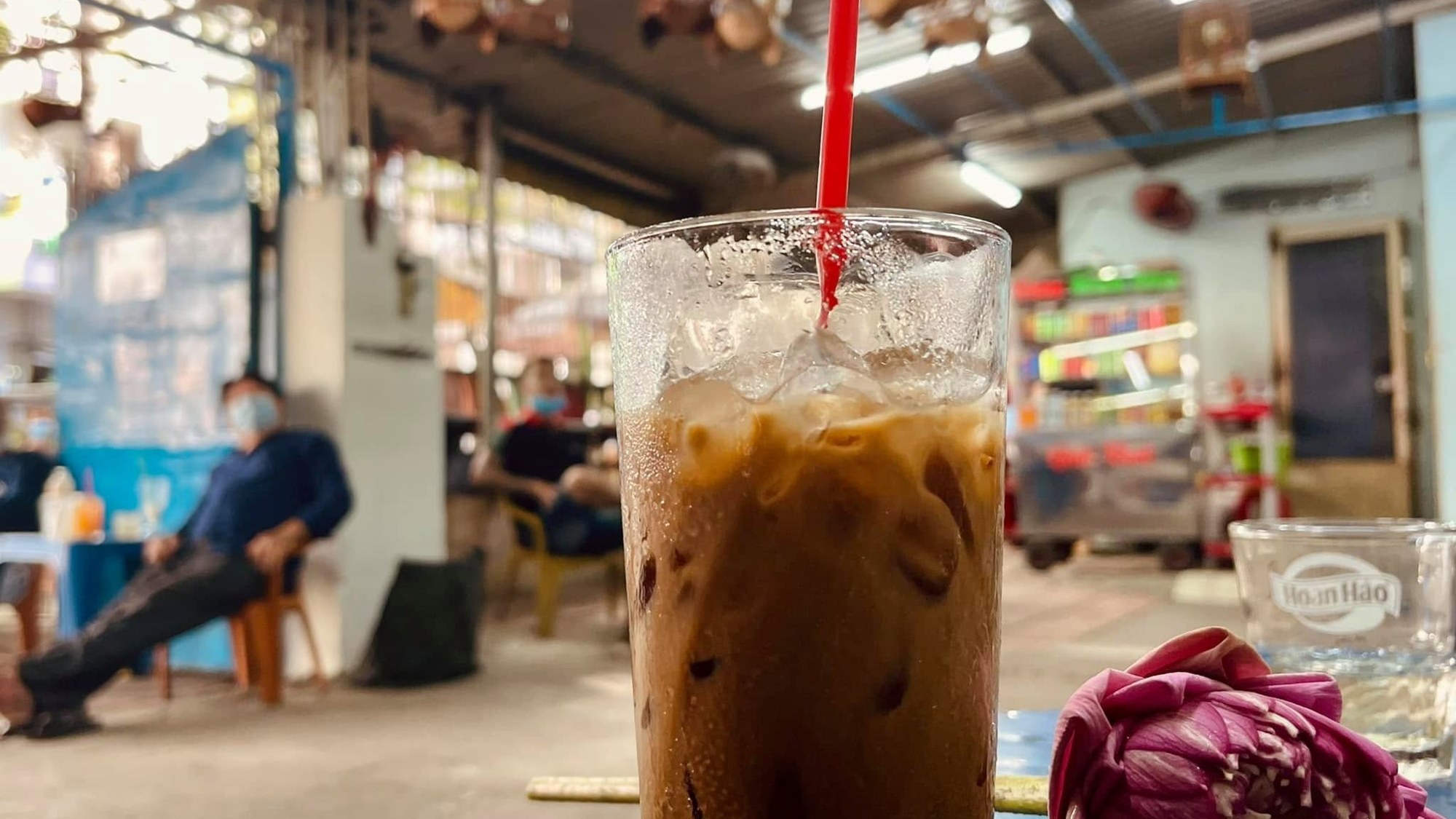 An iced coffee in a tall glass on a café table at a Saigon bird café, cages hanging from the roof above.