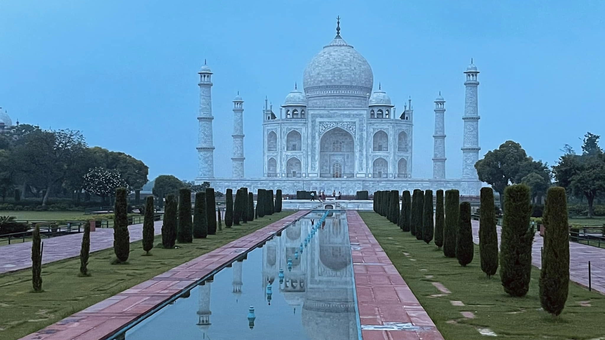 The Taj Mahal at dawn under a blue sky, reflected in the long garden pool between rows of cypress trees.