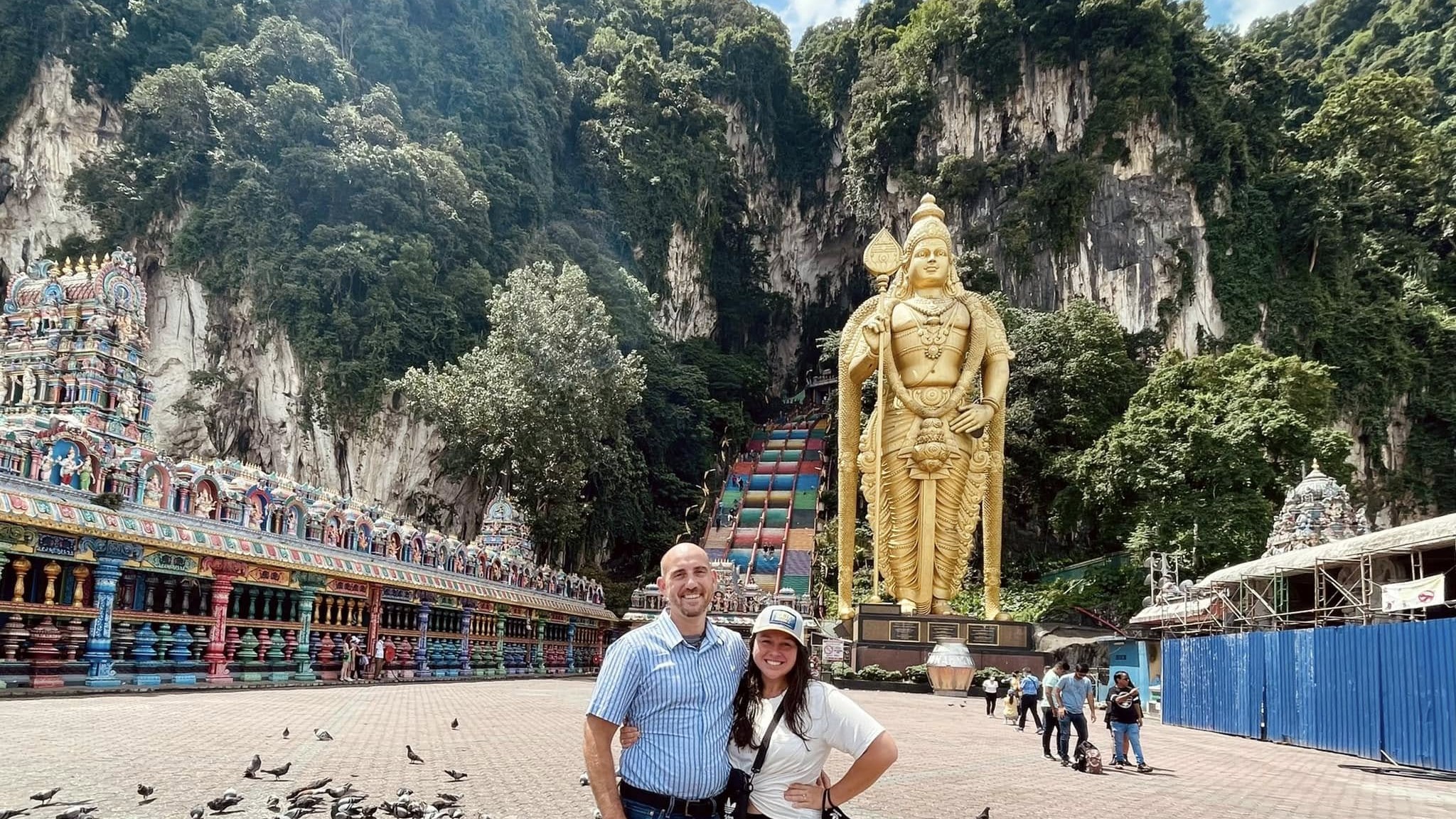 Nancy and Jay on the plaza at Batu Caves, the gold statue of Lord Murugan and the rainbow stairs behind them.