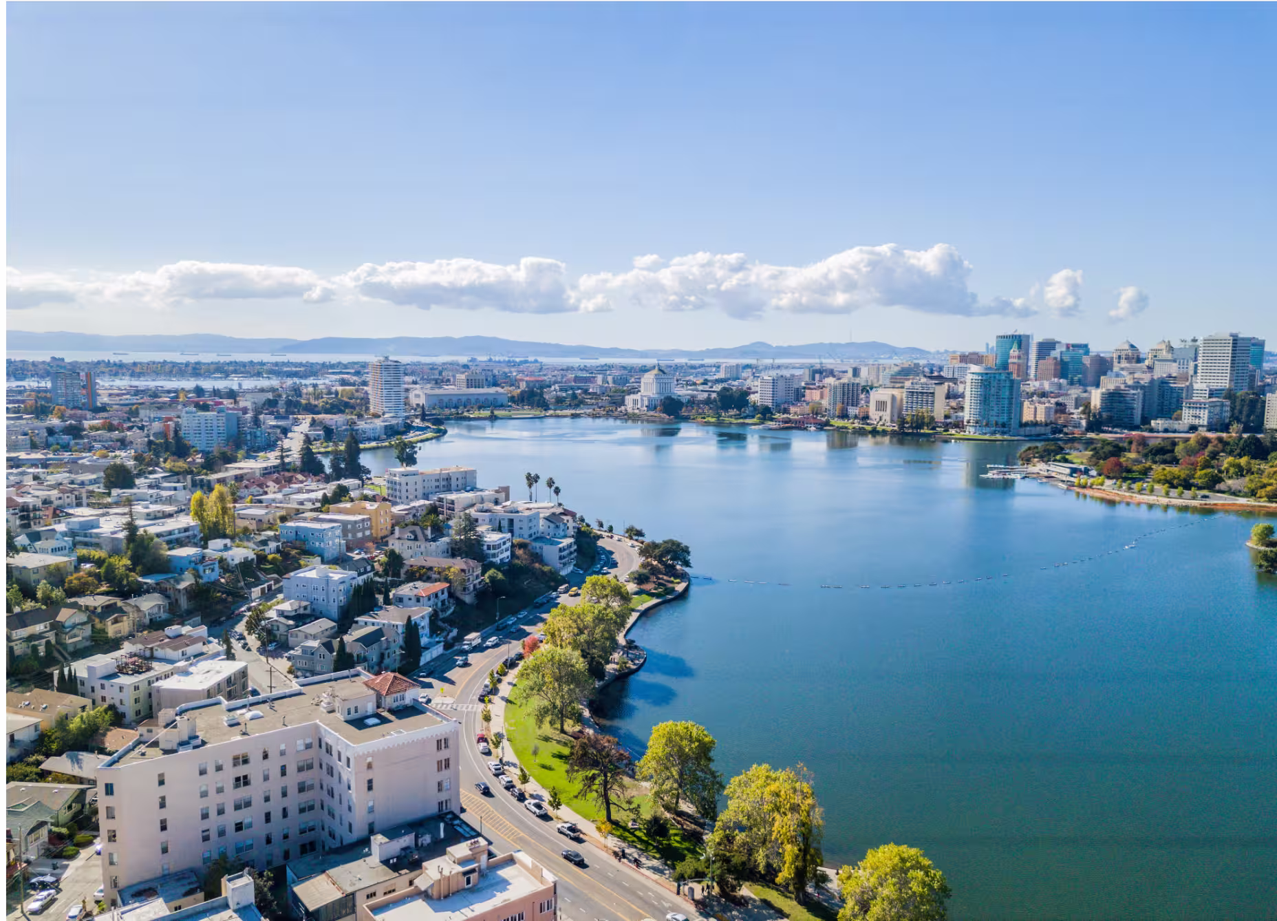 Aerial view of a city with a large lake, curved road, greenery, and buildings under a clear sky.