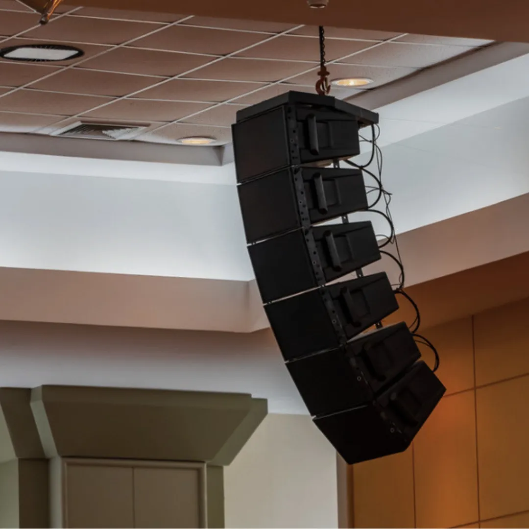 Flown line-array speaker cluster of six cabinets rigged from a ceiling chain hoist, angled toward the room below