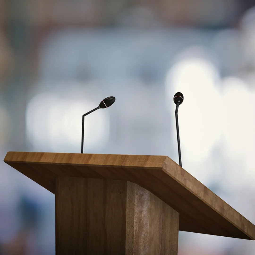 Wooden lectern with two black gooseneck microphones against a blurred, brightly lit background