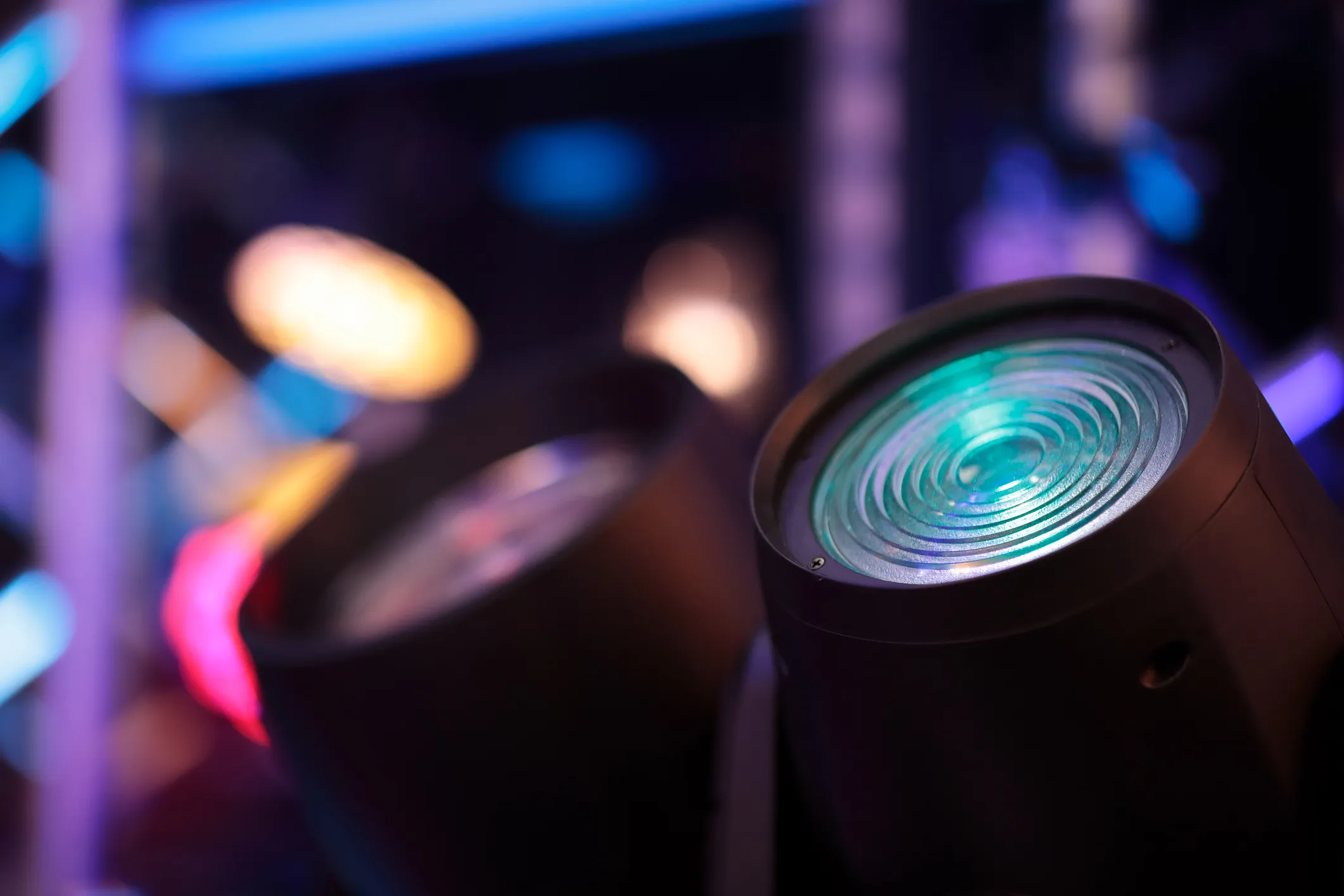 Close-up of stage moving-head light fixtures with a teal lens, blurred colorful stage lighting in the background