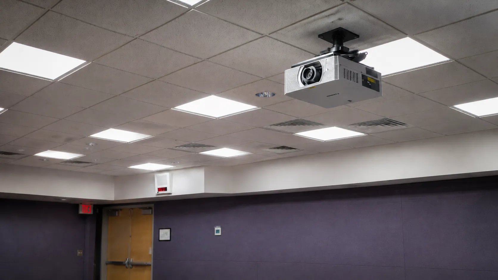 Canon ceiling-mounted projector installed in a drop ceiling above a room with purple acoustic-paneled walls and a wood door