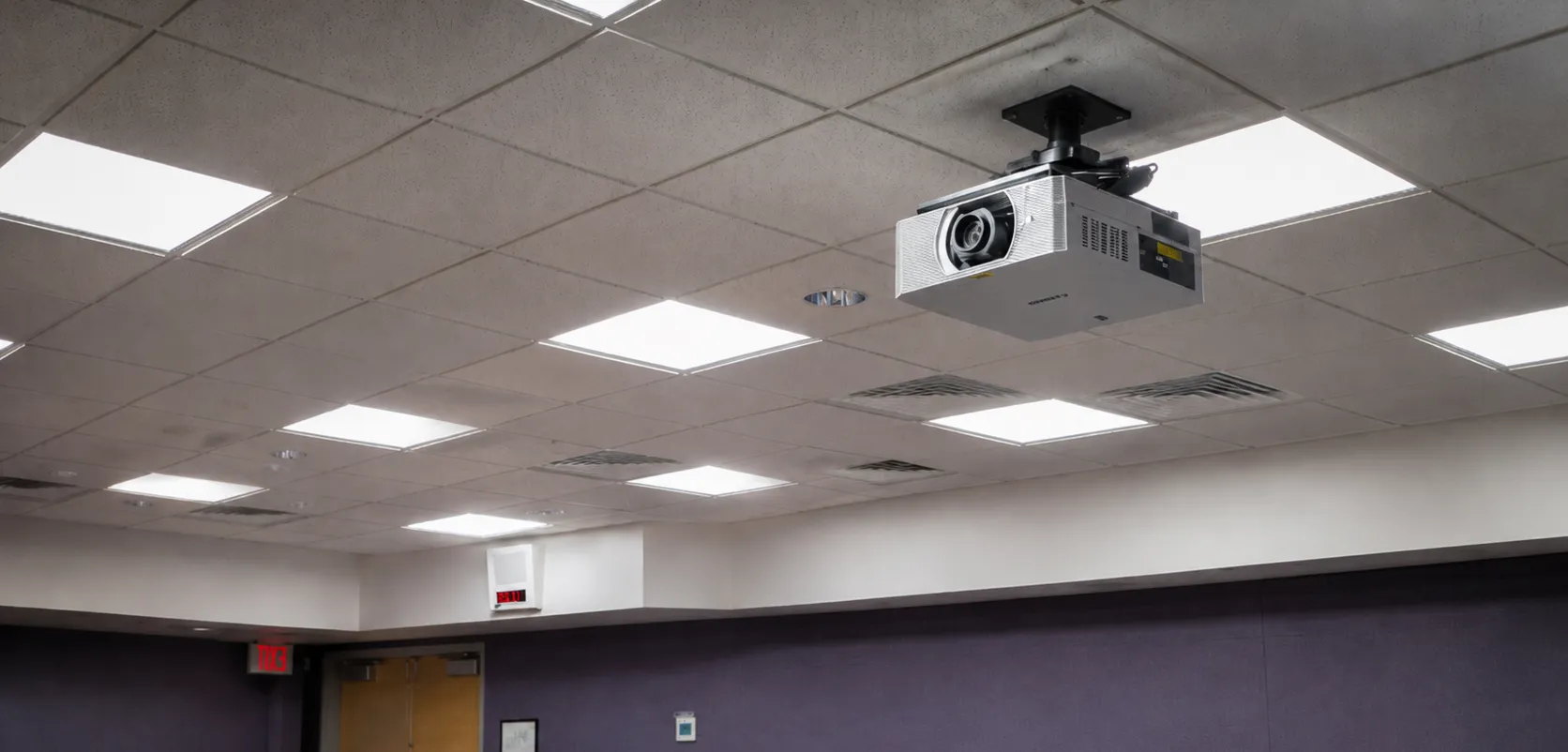 Canon ceiling-mounted projector installed in a drop ceiling above a room with purple acoustic-paneled walls and a wood door