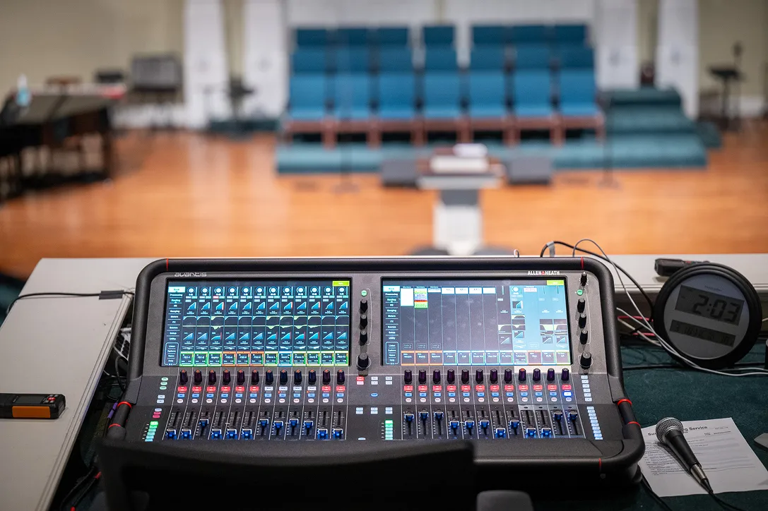 AV control desk at a church with an Allen & Heath Avantis digital mixer, an iMac running ProPresenter with slide graphics, and a MIDI fader controller, facing rows of blue auditorium seating