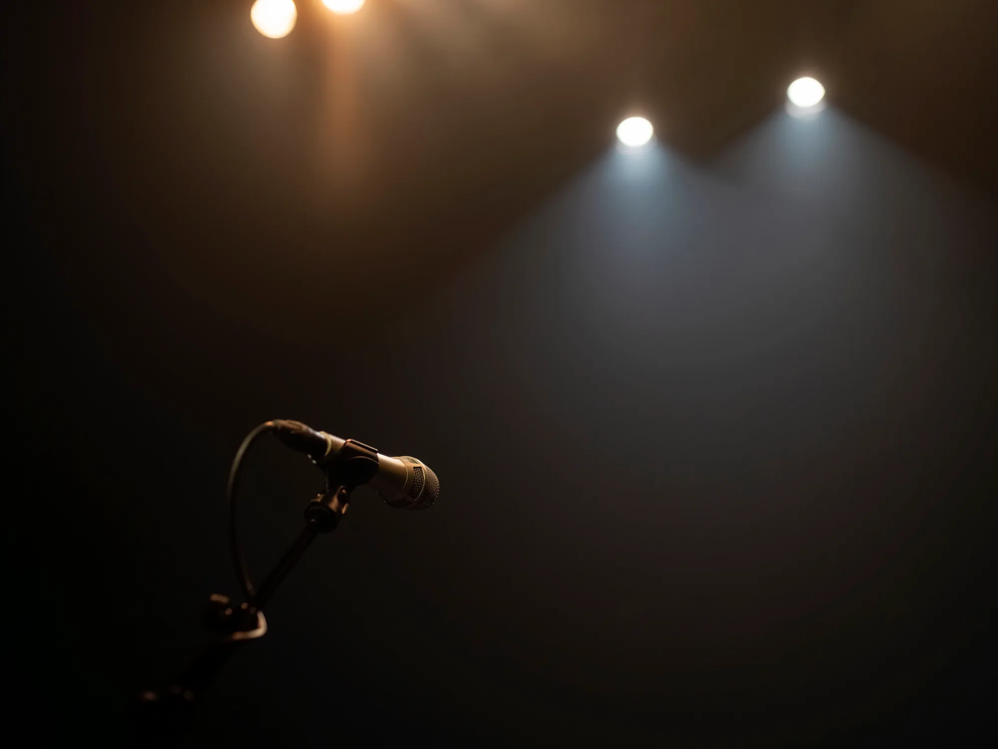 Silhouette of a stage microphone on a stand beneath hazy spotlight beams against a dark background