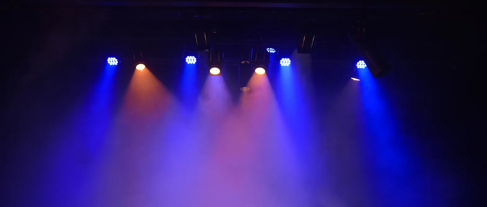 Wide banner of a stage lighting rig with blue and amber spotlight beams cutting through haze