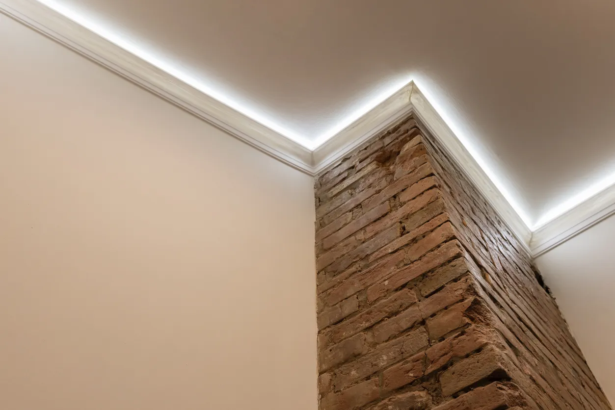 Warm LED cove lighting tracing crown molding above an exposed brick corner where two walls meet