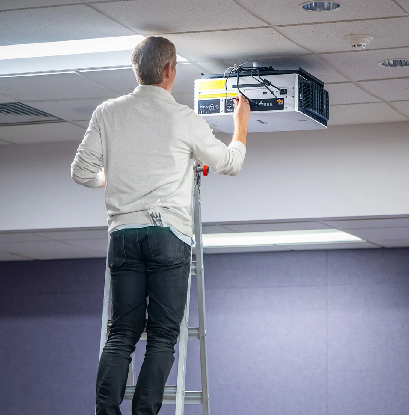 Technician viewed from behind on a ladder, adjusting cables on a ceiling-mounted projector in a room with purple acoustic wall panels