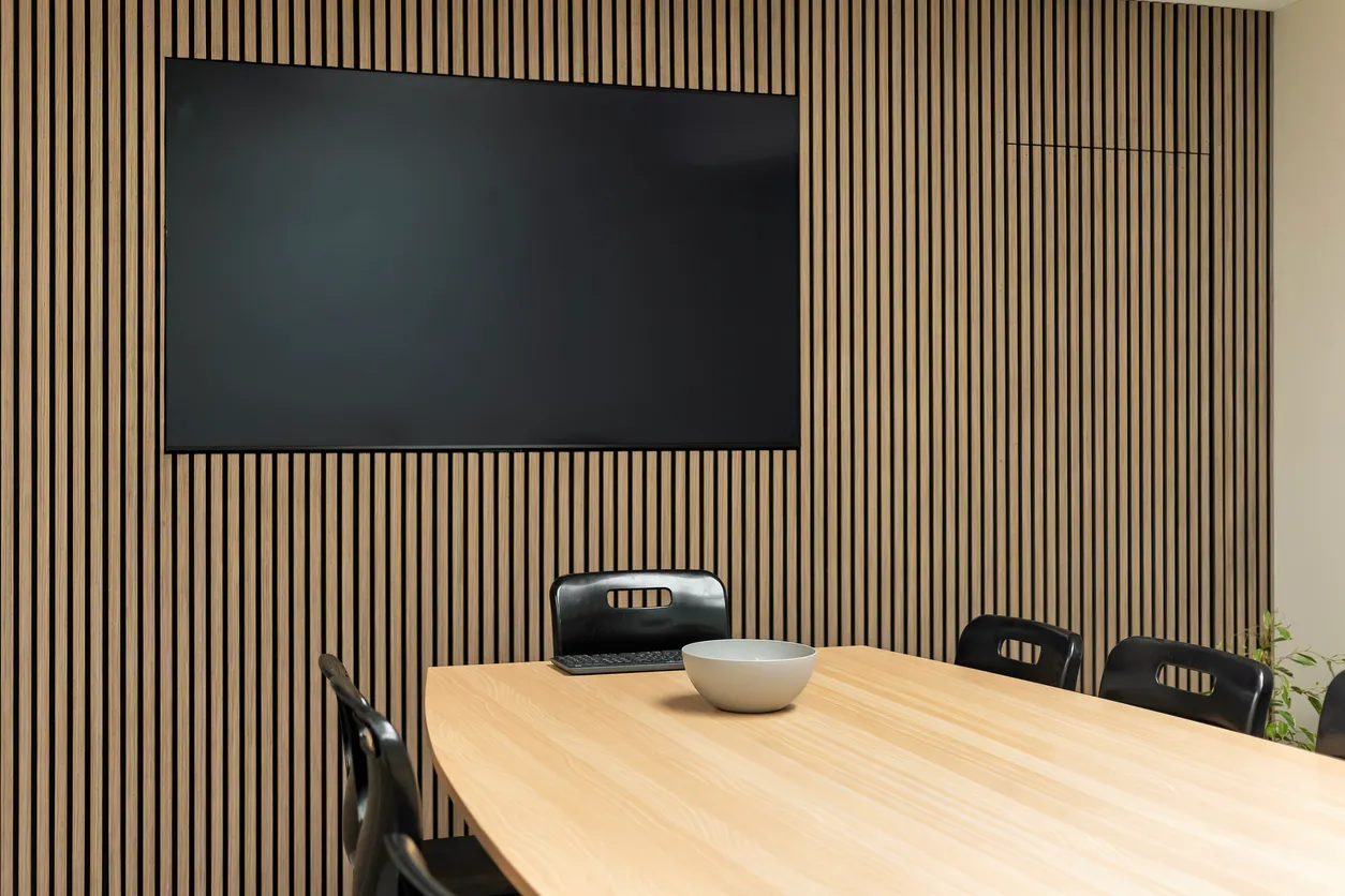 Conference room with a wall-mounted display on a vertical wood-slat wall, above a light wood table with a bowl and tablet remote